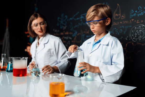 Teacher support schoolboy in laboratory they wear lab coat and glasses stand and experiment about science of chemistry in STEM class. Student tong solid down to yellow liquid in beaker. Erudition. - Photo, Image
