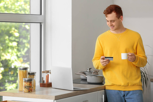 Przystojny młody człowiek z karty kredytowej i filiżanki kawy zakupy online w kuchni - Zdjęcie, obraz