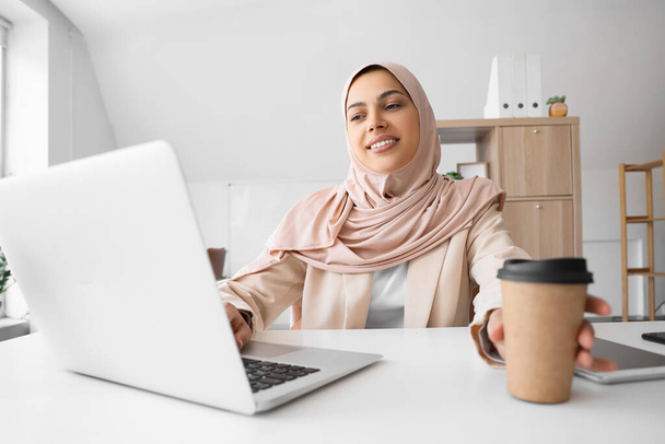 Όμορφη μουσουλμάνα γυναίκα προγραμματίστρια με φλιτζάνι καφέ εργάζεται στο γραφείο - Φωτογραφία, εικόνα