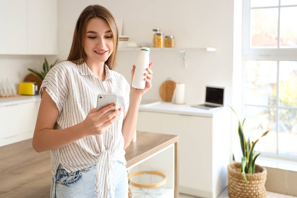 Nuori nainen pullon maukasta jogurttia käyttäen matkapuhelinta keittiössä - Valokuva, kuva