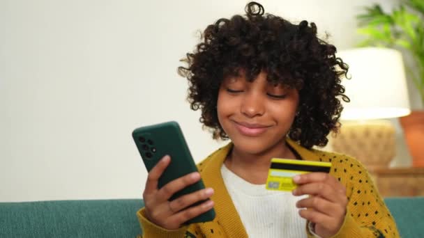African American woman shopping online holding smartphone paying with gold credit card. Girl sitting at home buying on Internet enter credit card details. Online shopping ecommerce delivery service - 映像、動画