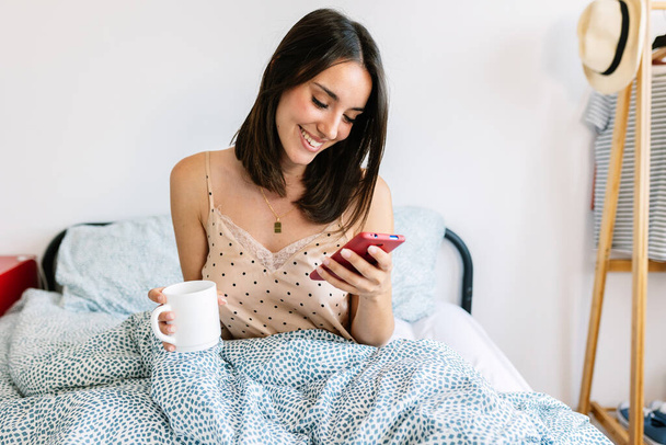 Νεαρή όμορφη γυναίκα που χρησιμοποιεί το κινητό τηλέφωνο πίνοντας καφέ το πρωί μέσα στο κρεβάτι. - Φωτογραφία, εικόνα