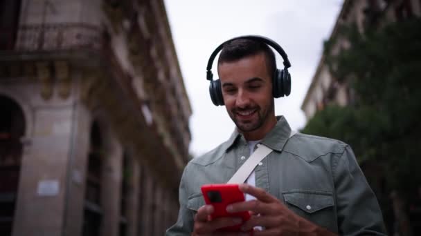 Jovem homem atraente com fones de ouvido andando gosta de ouvir música acenando com a cabeça olhando telefone celular na rua na cidade europeia de Madrid. Passeio turístico masculino feliz usando o turismo de viagens ao ar livre de células vermelhas - Filmagem, Vídeo