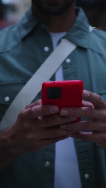 Вертикальний фокус руки молодий кавказький чоловік ходить за допомогою червоного мобільного телефону щасливий на вулиці в сутінках в європейському місті. Веселий чоловічий турист насолоджується прогулянкою, переглядом мобільного друку досвіду подорожей на відкритому повітрі - Кадри, відео