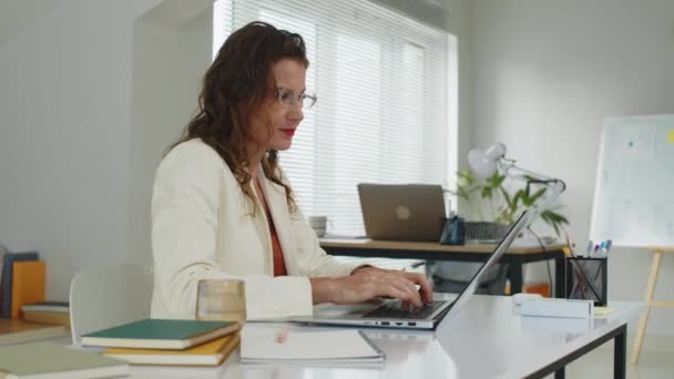 Vista lateral de empleada femenina pensativa de etnia caucásica que comprueba informe en el ordenador portátil inalámbrico en la oficina - Metraje, vídeo