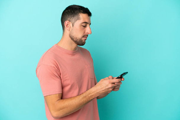 Giovane uomo caucasico bello isolato su sfondo blu invio di un messaggio o e-mail con il cellulare - Foto, immagini