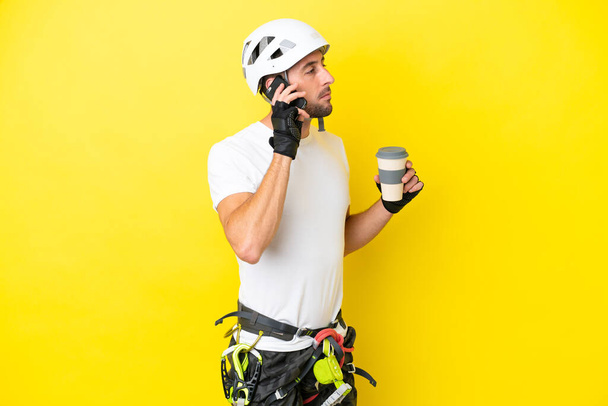 Young rock- climber man isolated on yellow background holding coffee to take away and a mobile - Photo, Image