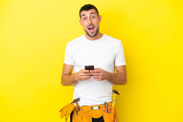 jonge elektricien blanke man geïsoleerd op gele achtergrond verrast en het verzenden van een bericht - Foto, afbeelding