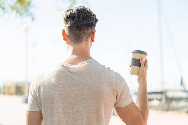 Young handsome man holding a take away coffee at outdoors in back position - Photo, Image