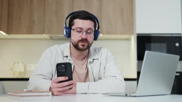 Relaxed man in wireless headphones looking at smartphone screen while shaking head and singing along to music. Careless freelancer sitting next to working computer and enjoying favourite song. - Footage, Video
