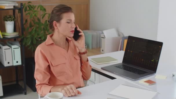 Střední záběr zaneprázdněné ženy podnikatel mluví o finanční graf na mobilním telefonu sedí u stolu s bezdrátovým notebookem - Záběry, video