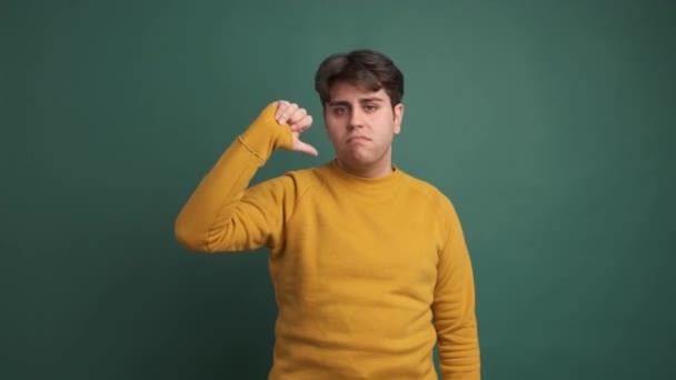 Portrait of displeased Hispanic young man in yellow outfit showing thumb down and shaking head while looking at camera against green background - Footage, Video