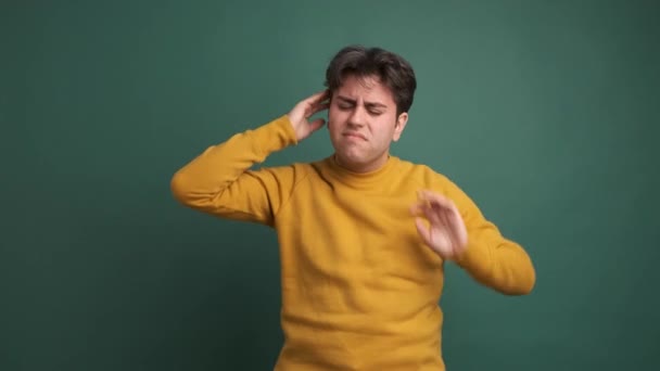 Hombre joven hispano pensativo en traje amarillo masajeando la cabeza que sufre de dolor de cabeza sobre fondo verde - Metraje, vídeo