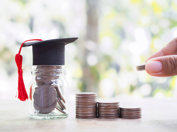 Γυάλινο μπουκάλι με καπέλο αποφοίτησης και χέρι βάζοντας κέρμα σε στοίβα νομισμάτων. Η έννοια της εξοικονόμησης χρημάτων για την εκπαίδευση, φοιτητικό δάνειο, υποτροφία, δίδακτρα στο μέλλον - Φωτογραφία, εικόνα