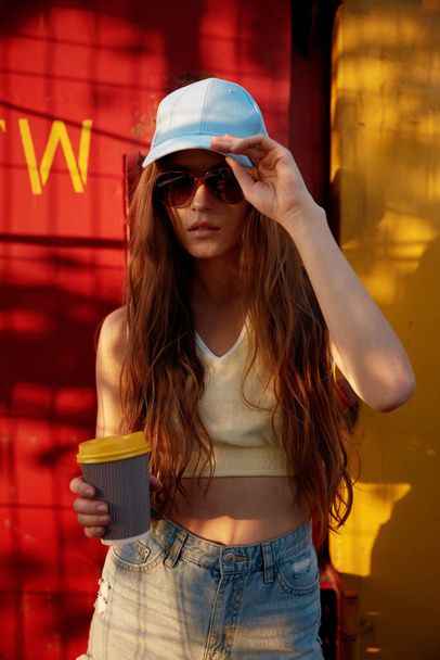 Cool hipster woman skateboarder wearing casual trendy outfit drinking coffee standing in shadow of tree. Urban skateboard park and fashionable teenage style - Photo, Image