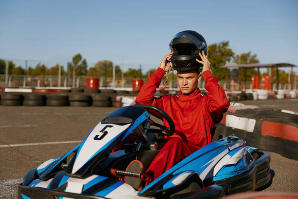 Handsome man driver wearing uniform taking off protective helmet while sitting at kart race car. Extreme go-kart training on race track - Photo, Image
