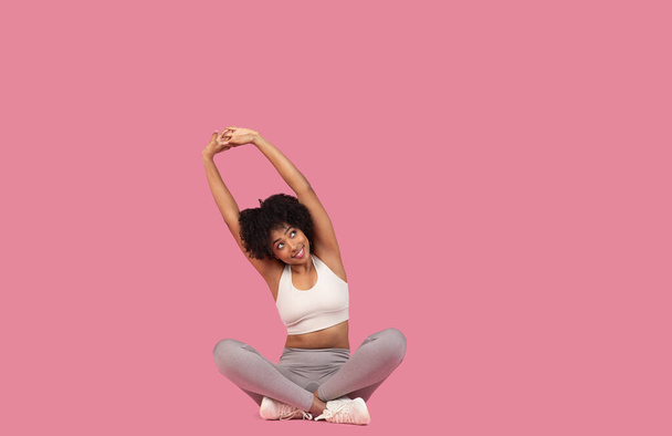 Happy Afro-Amerikaanse vrouw in witte sport top en grijze leggings doen stretch met armen boven het hoofd, zitten op roze achtergrond, vrije ruimte - Foto, afbeelding