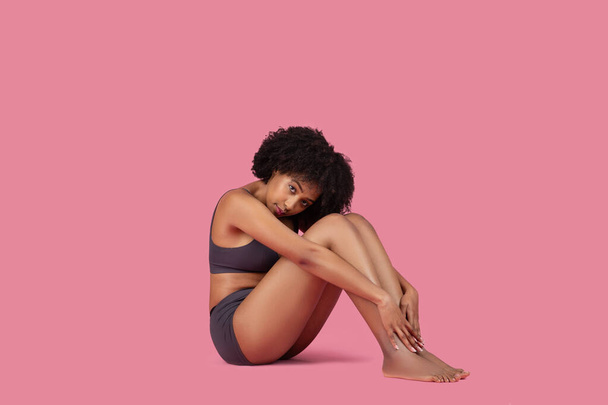 Pensive fiatal afro-amerikai nő sportruházat vagy fehérnemű ül, és megérinti a lábát rózsaszín háttér, nézi a kamera, szabad hely - Fotó, kép