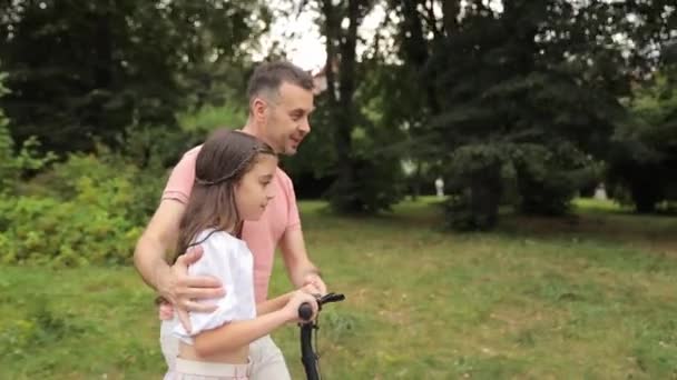 A man is pushing a little girl on a scooter through a park, surrounded by lush green grass, trees, and a peaceful natural landscape. They are enjoying leisure time outdoors - Footage, Video