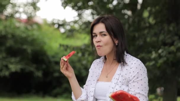 Een vrouw geniet van een stuk watermeloen onder een boom in het park. Haar gelukkige gebaar laat zien hoezeer ze geniet van het verfrissende fruit terwijl ze omringd wordt door groene bomen in het park. - Video
