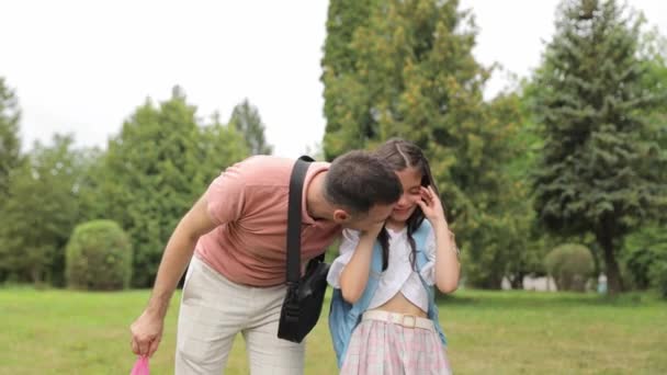 A man and a little daughter are standing near a tree in the park, sharing a happy moment in nature. The man gestures towards the plant as they enjoy leisure and entertainment together - Footage, Video