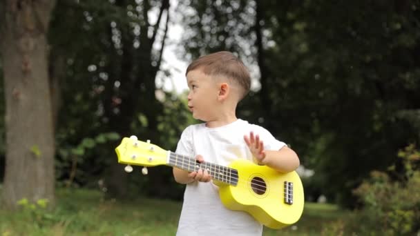 Un niño músico está feliz de tocar el ukelele amarillo en el césped del parque, haciendo buena música en el parque. - Imágenes, Vídeo