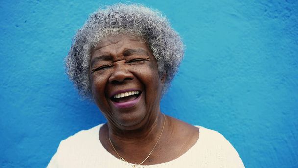 Одна счастливая пожилая афроамериканка с седыми волосами смеется и улыбается на фоне синей стены. Портрет радостного латинского пожилого человека - Фото, изображение