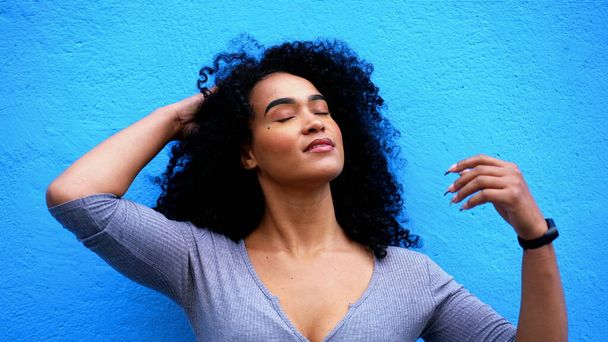 Uma jovem mulher negra atraente sorrindo para a câmera em ambiente urbano em pé na parede azul. 20s Africano descendente menina adulta ajustando cabelo encaracolado sentindo-se capacitado e confiante - Foto, Imagem
