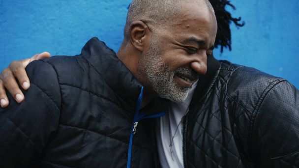 Afroamerikanisch erwachsener Sohn umarmt Vater mittleren Alters in aufrichtigem fürsorglichen Moment zwischen Eltern und Sprösslingen, setzt Stirn zusammen und Emblem auf blauem städtischen Hintergrund - Foto, Bild