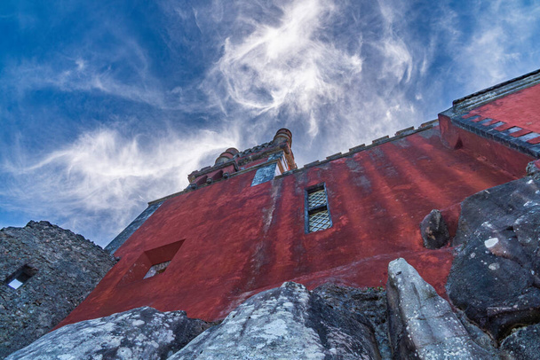 Niedrige Ansicht eines Teils einer roten Wand des Pena-Palastes auf einigen großen Felsen unter blauem Himmel mit weißen Spinnwebenwolken, die von der Sonne hinterleuchtet werden. Sintra. Portugal. - Foto, Bild