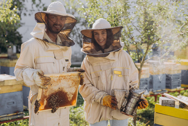 Coppia di felici apicoltori sorridenti che lavorano con gli strumenti di apicoltura vicino all'alveare nella fattoria delle api - Foto, immagini