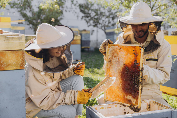 蜂の農場の養蜂場の近くで養蜂用具を使用している幸せな微笑みの養蜂家のカップル - 写真・画像