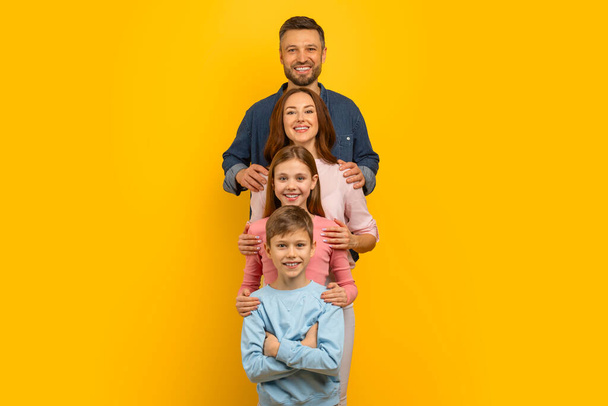 Jovem família feliz, mãe, pai, filha e filho abraçando enquanto em pé na fila, pais caucasianos alegres e seus filhos se unindo, posando no fundo do estúdio amarelo, espaço de cópia - Foto, Imagem