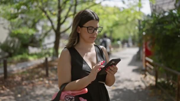 Красива іспаномовна жінка, використовуючи мобільний телефон, радісно посміхається під час прогулянки на традиційних вулицях Кіото з її милими окулярами на - Кадри, відео