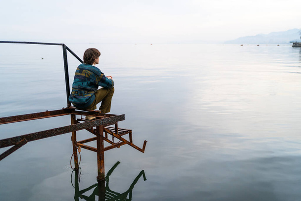 Homme pensif assis seul sur une jetée altérée par la mer tranquille au crépuscule, horizon s'étendant au-delà. - Photo, image