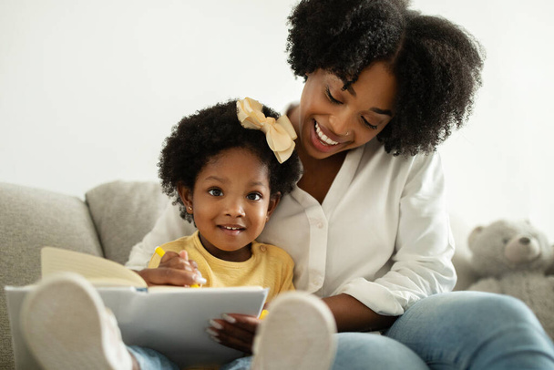 Rozwój dziecka. Całkiem młoda czarna mamusia czyta książkę ze swoją słodką córeczką w domu, szczęśliwa Afroamerykanka spędza czas z małym dzieckiem, cieszy się urlopem macierzyńskim, zbliżeniem - Zdjęcie, obraz