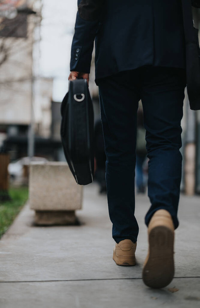 Κοντινό πλάνο ενός επαγγελματία με πόδια που περπατούν έξω, κουβαλώντας ένα δερμάτινο χαρτοφύλακα σε αστικό περιβάλλον. - Φωτογραφία, εικόνα