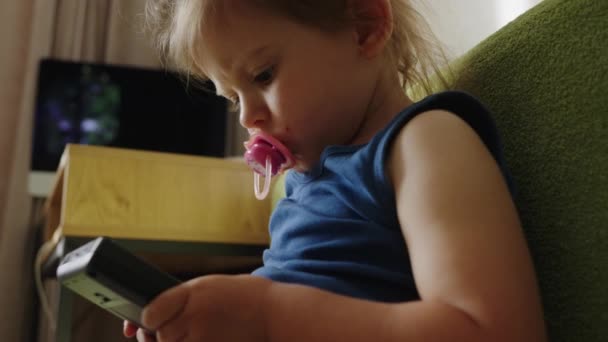 Piccolo bambino seduto sul divano verde succhiare un ciuccio che tiene un telefono nero gioco. - Filmati, video