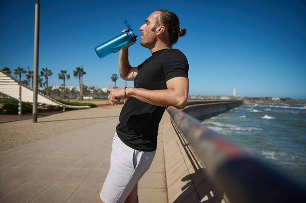 Glücklich entschlossener, athletischer Mann in schwarzem T-Shirt und grauer Sporthose, trinkt Wasser aus Sportflasche, ruht sich nach dem morgendlichen Joggen aus, steht an einer Brücke an der Atlantikpromenade - Foto, Bild
