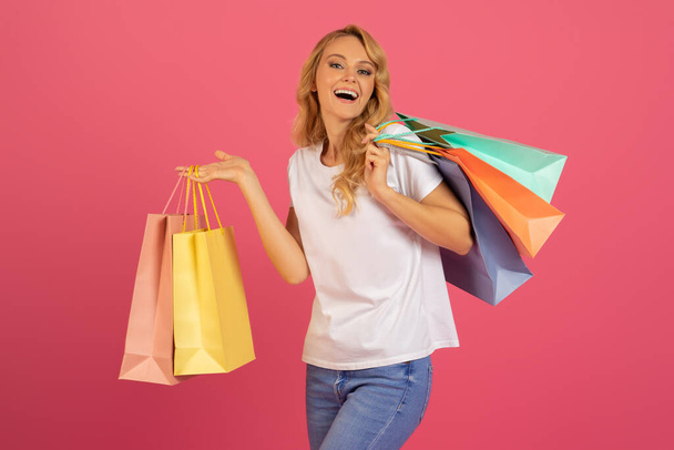 Kausikauppias. Kaunis eurooppalainen blondi nainen kuljettaa ostoskasseja ja ilmaista onnea yli suuri myynti, seisoo ostot studiossa asetelma vaaleanpunainen tausta. Alennuksen edistäminen - Valokuva, kuva