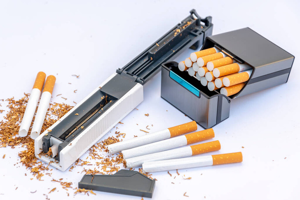 Plnicí stroj na cigaretová pouzdra s tabákem, roztroušené prázdné cigarety a hromádka tabáku na bílém pozadí, pouzdro na cigarety s domácími cigaretami. - Fotografie, Obrázek