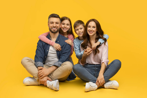 Happy young family of four sitting on floor and embracing, cheerful loving parents posing together with their son and daughter on yellow studio background, looking at camera, copy space - Photo, Image