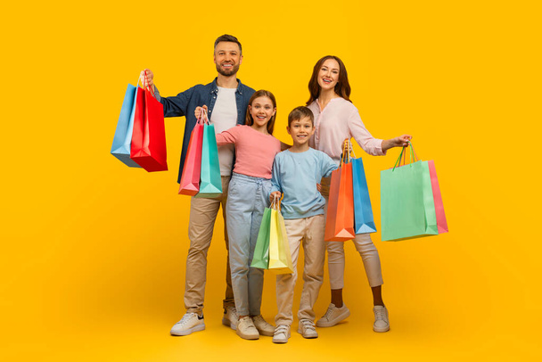 Family Shopping. Joyful Young Parents And Their Kids Holding Bright Shopper Bags And Having Fun Together While Standing On Yellow Background, Cheerful Mom, Dad And Children Enjoying Seasonal Sales - Photo, Image