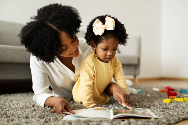 Любить молодую африканскую американскую мать, читающую книгу со своим ребенком, милой маленькой девочкой. Мама и дочь лежат на полу дома, проверяют красочные картинки на плэйбуке, копируют пространство - Фото, изображение