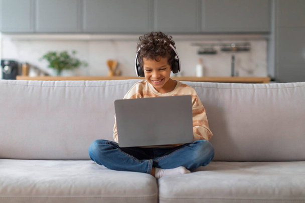 Boldog fekete tinédzser fiú visel fejhallgató segítségével laptop, miközben ül keresztbe lábú kanapén otthon, mosolygós afro-amerikai férfi gyerek elmerül az interaktív oktatási tevékenység vagy online játék - Fotó, kép