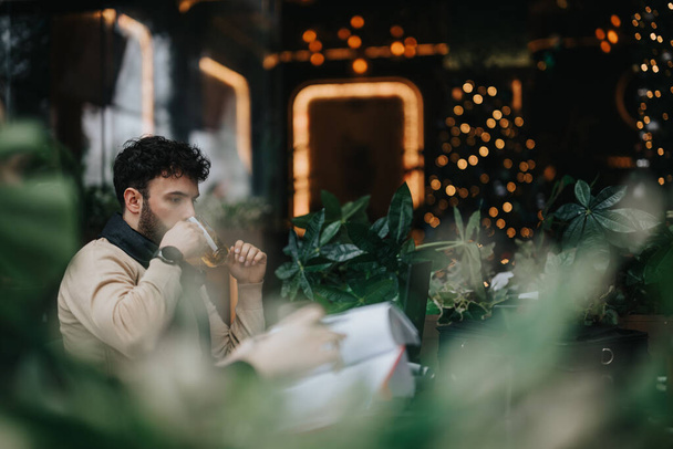 Ein junger Mann nippt intensiv an einem Drink und konzentriert sich auf seine Arbeit im Ambiente eines trendigen städtischen Cafés, umgeben von Bokeh-Lichtern. - Foto, Bild