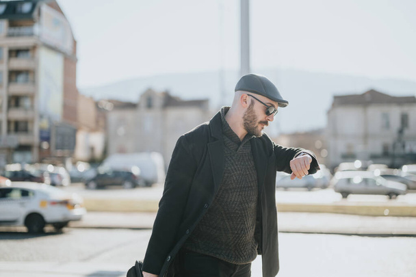 Elegante hombre de negocios caminando y comprobando el tiempo en reloj de pulsera en un entorno urbano soleado. - Foto, Imagen