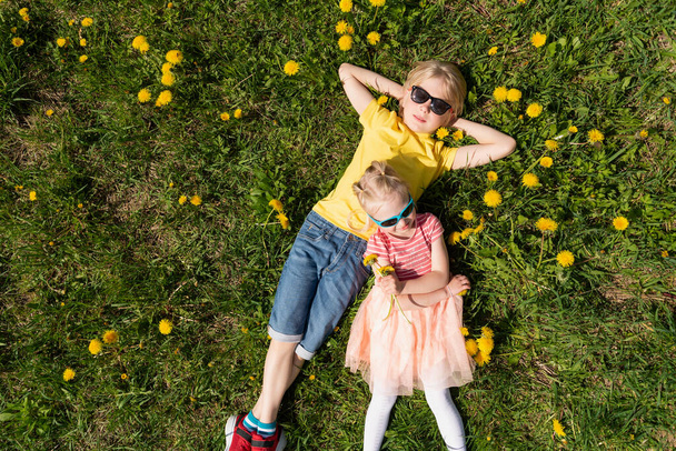 Fratellino e sorella crogiolarsi al sole nei meadow.Two bambini biondi in occhiali da sole sdraiati sul campo di denti di leone. - Foto, immagini