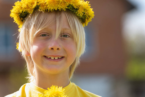 Ritratto di ragazzo biondo sorridente con corona di denti di leone e bouquet di fiori. Giornata di sole. Sfondo sfocato - Foto, immagini