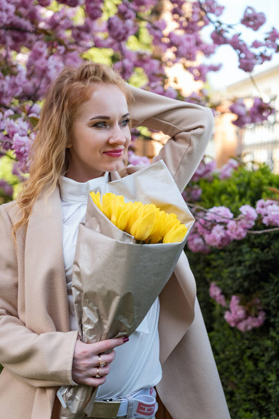 Молода приваблива жінка тримає в парку букет жовтих тюльпанів. Жінка з квітами на вулиці чекає когось. Вертикальна рамка. - Фото, зображення